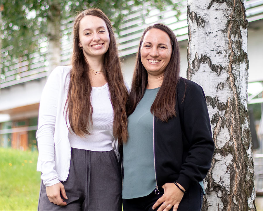 Das Bild zeigt eine junge, freundliche Frau mit schulterlangem braunem Haar, die entspannt in einem hellen, modernen Sessel sitzt. Sie trägt ein hellblaues Jeanshemd und hat ein silbernes Notebook auf den Oberschenkeln abgelegt. In der rechten Hand hält sie ein Smartphone ans Ohr und führt offenbar ein angenehmes Telefonat – ihr Gesichtsausdruck ist offen und lächelnd. Im Hintergrund ist ein minimalistischer Wohnraum mit hellen Farben, einem kleinen Regal und einer grünen Topfpflanze auf einem Tisch zu sehen. Die natürliche Beleuchtung sorgt für eine warme, einladende Atmosphäre.